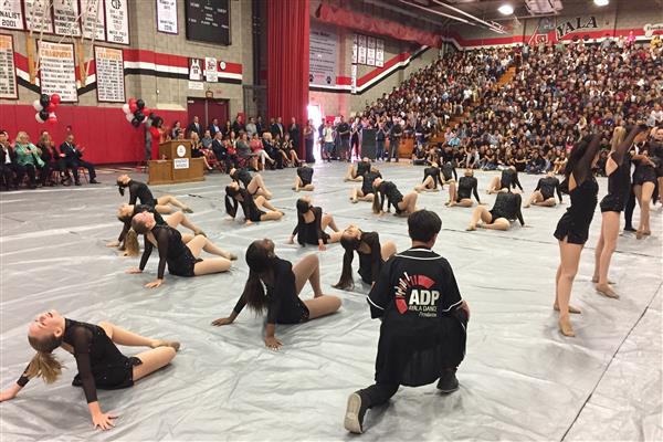 Several female high school students on a gym floor, in a dance pose. People watching from bleachers and chairs.